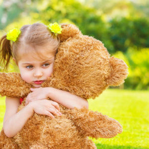 Little cute sad girl holding in hands brown teddy bear, upset child spending time outdoors in spring time